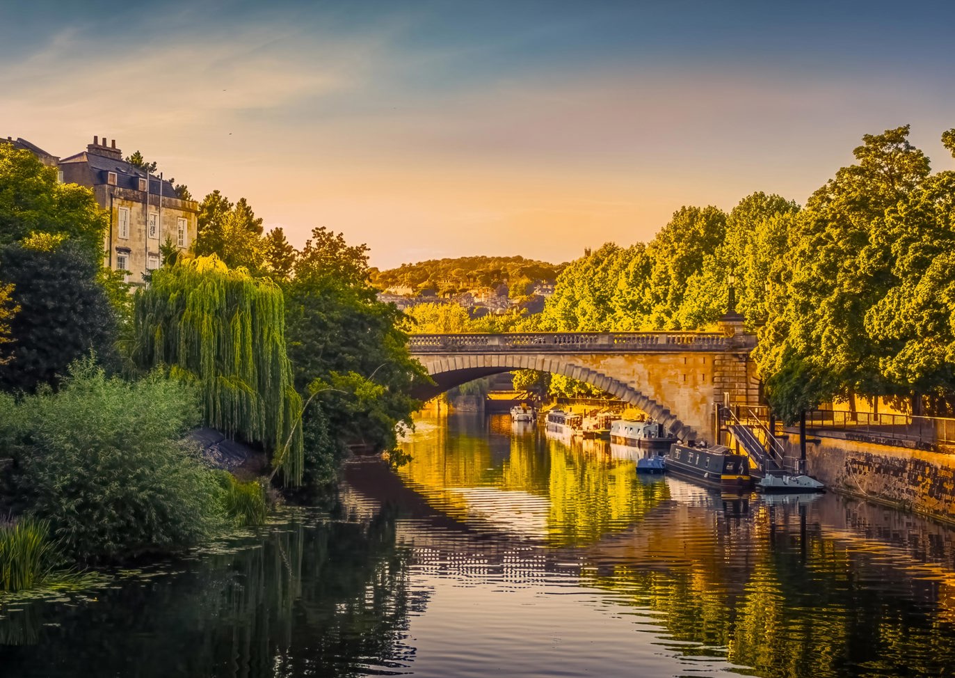 Beautiful view of the River Avon in Bath with a stone bridge and boats along the riverside at golden hour, Near Brokerswood Holiday Park.