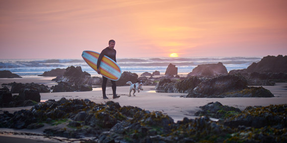 Mature surfer walking with surfboard and dog at sunset near Praa Sand Holiday Park