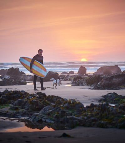 Mature surfer walking with surfboard and dog at sunset near Praa Sand Holiday Park