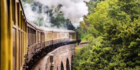 Steam train traveling across a stone viaduct surrounded by lush green trees, with smoke billowing from its engine