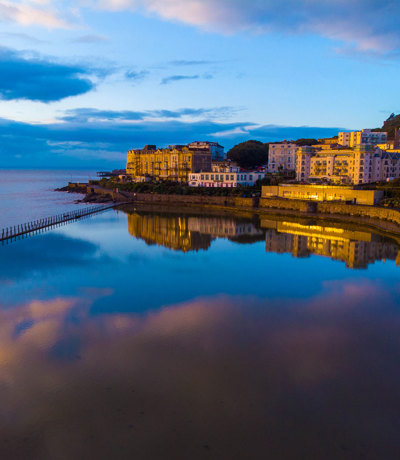 Coastal view of Weston-super-Mare at sunset golden hour, buildings reflecting on the calm water, Near Brokerswood Holiday Park.