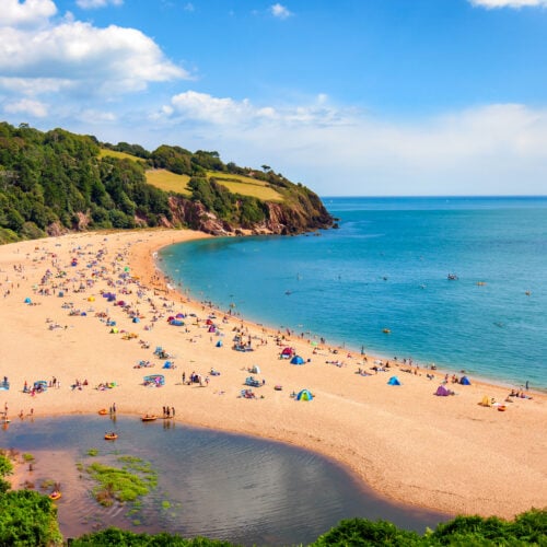 A sandy beach with a bay, bright blue waters, and holiday makers making the most of the sunshine.