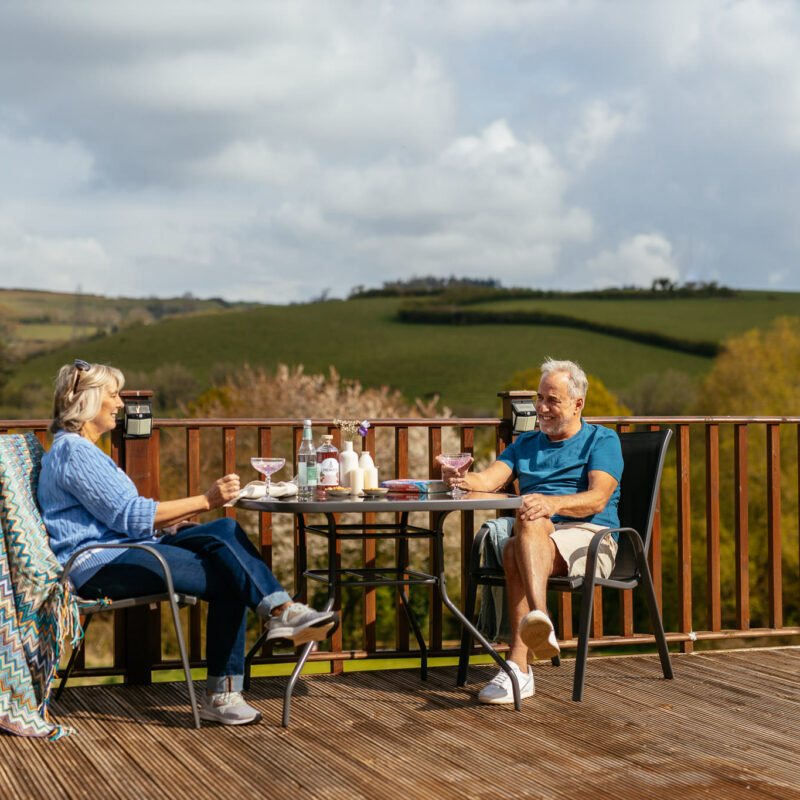 Older couple smiling, sitting at a table sipping on drinks, overlooking the Devon hills, Devon, England.